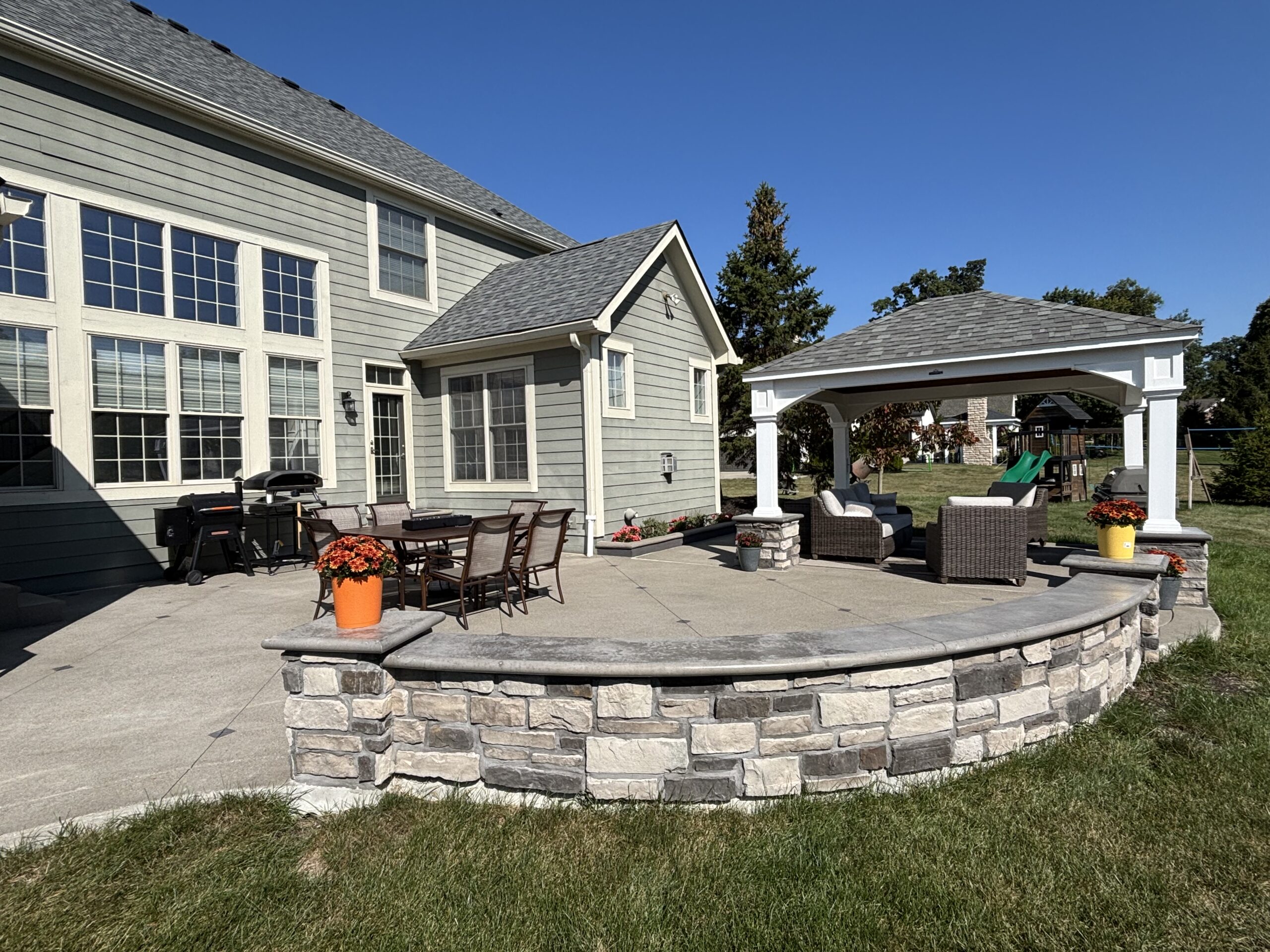 Backyard patio with a gray house, large windows, a stone retaining wall, orange potted flowers, and outdoor seating under a covered area.
