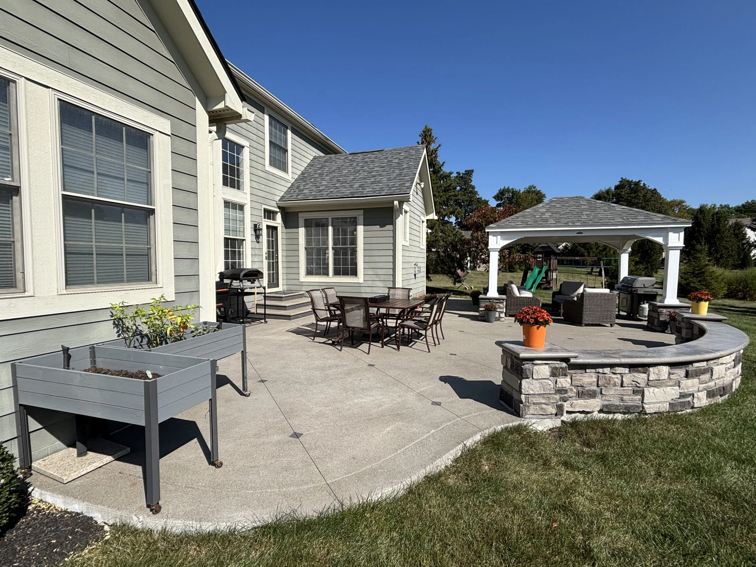 Backyard patio of a gray house with large windows, outdoor dining set, grill, and a stone fire pit; a white pergola covers part of the yard under a clear blue sky.