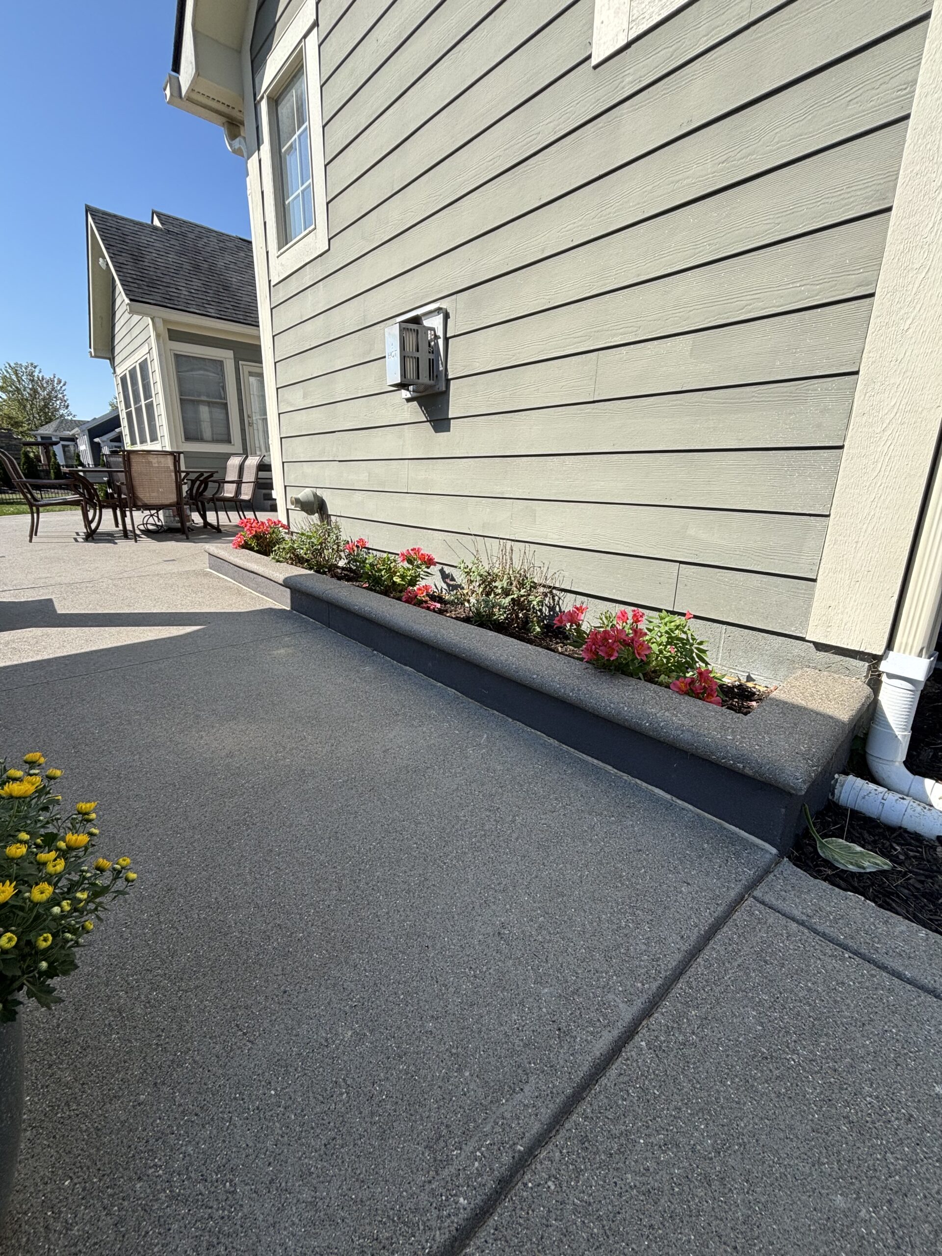 Patio area along a gray-laminate siding house with outdoor dining table and chairs, and a flower bed of pink blooms beside a concrete path.