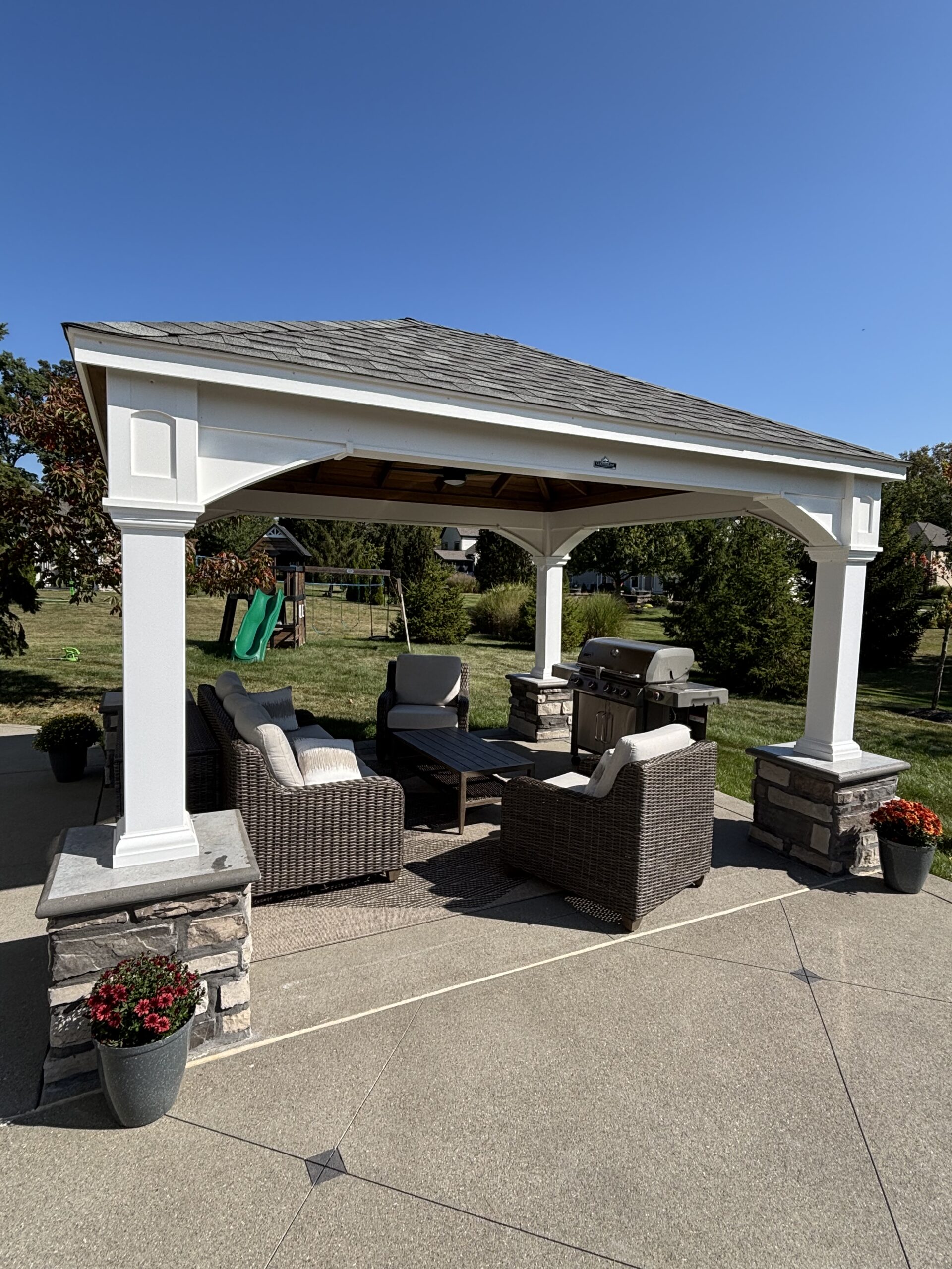 White outdoor gazebo with stone pillars and a seating area under a wooden roof, grill nearby, on a sunny day.