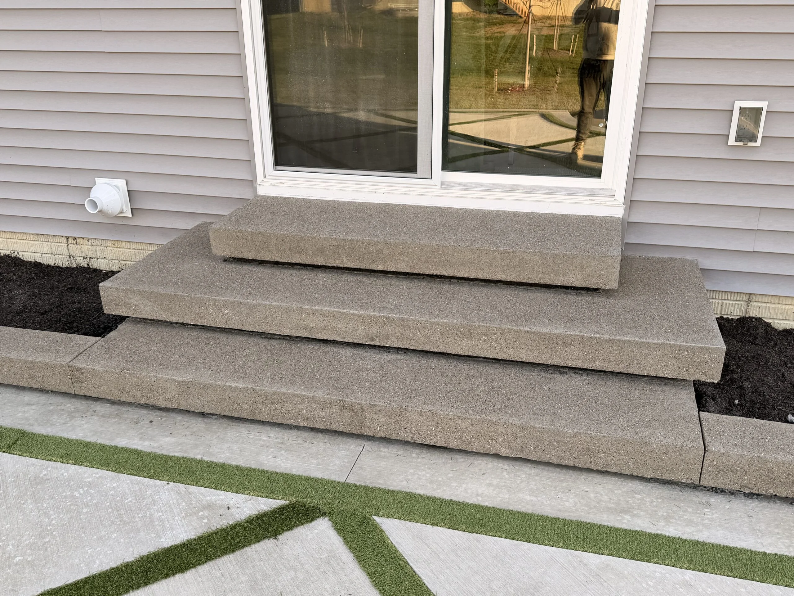 Concrete steps leading up to sliding glass door on a house with gray siding; exterior vent to the left and a small wall light to the right.