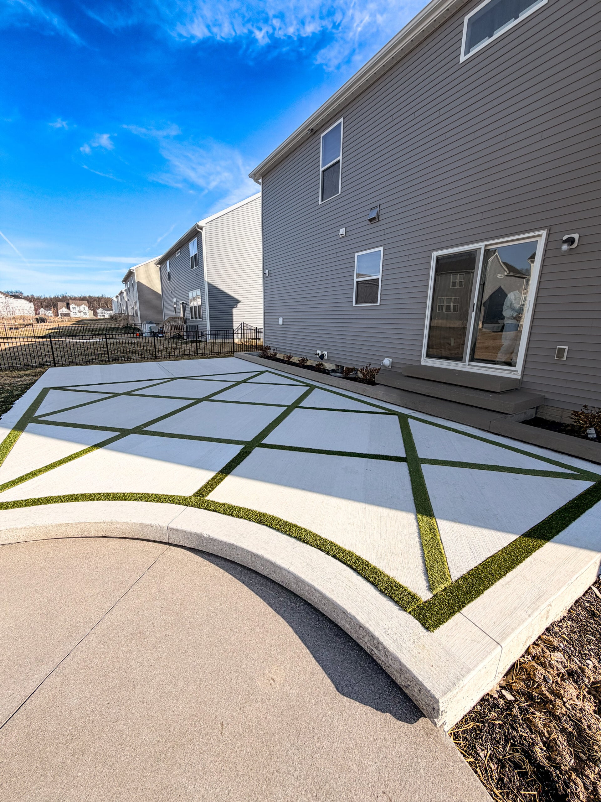 Modern townhouse row with gray siding and multiple windows beside a geometric concrete patio with green turf inlays under a blue sky