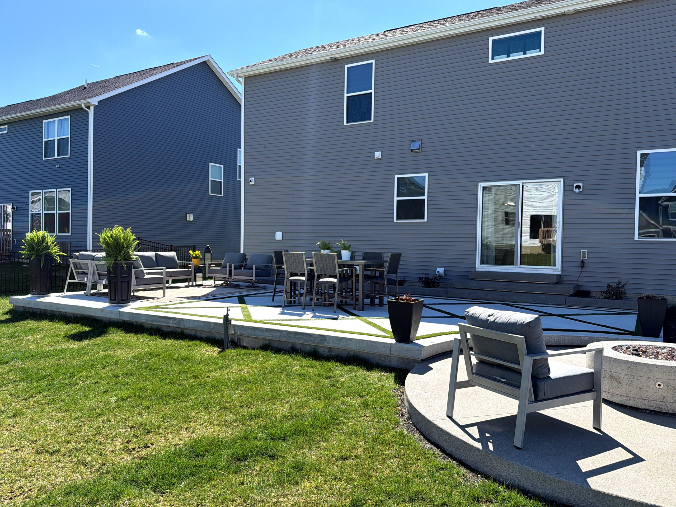 Patio with gray outdoor seating and a dining table, plants in planters, in front of two blue-gray suburban houses.