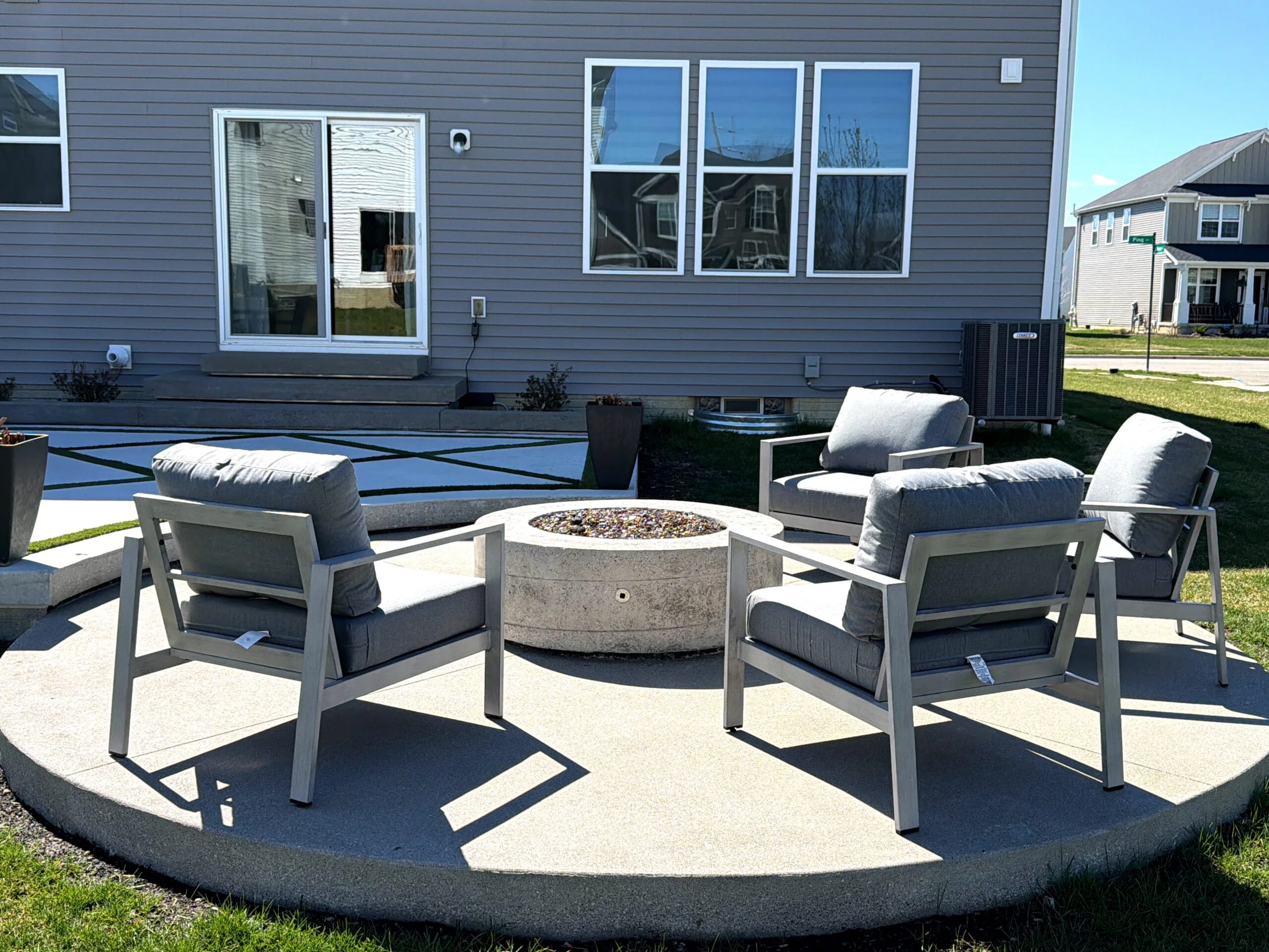 Backyard patio with a circular concrete fire pit surrounded by gray cushioned chairs in front of a gray house with large windows.