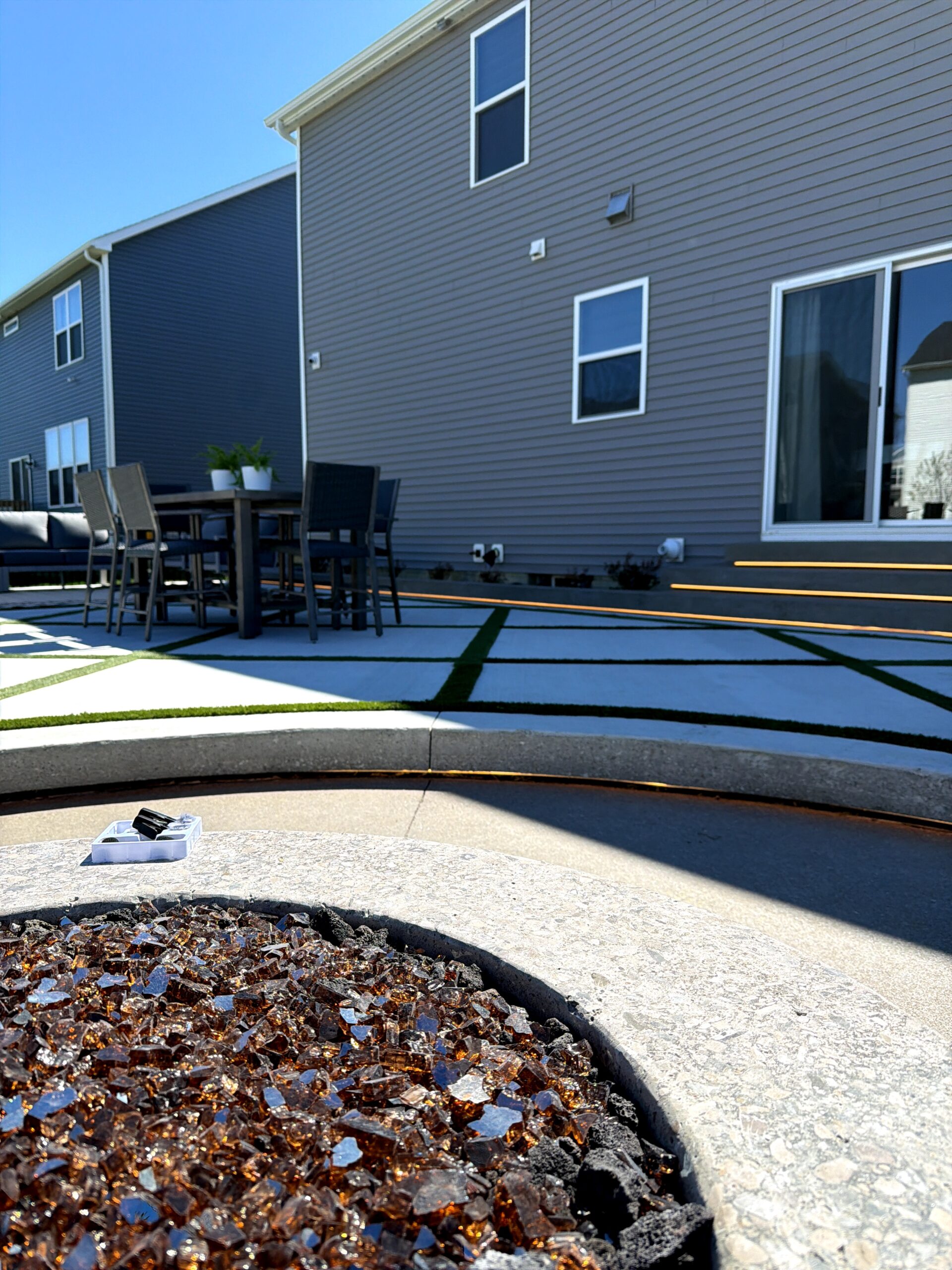 Backyard patio of a modern townhouse with a wooden table, chairs, and two potted plants, gray siding houses in the background.