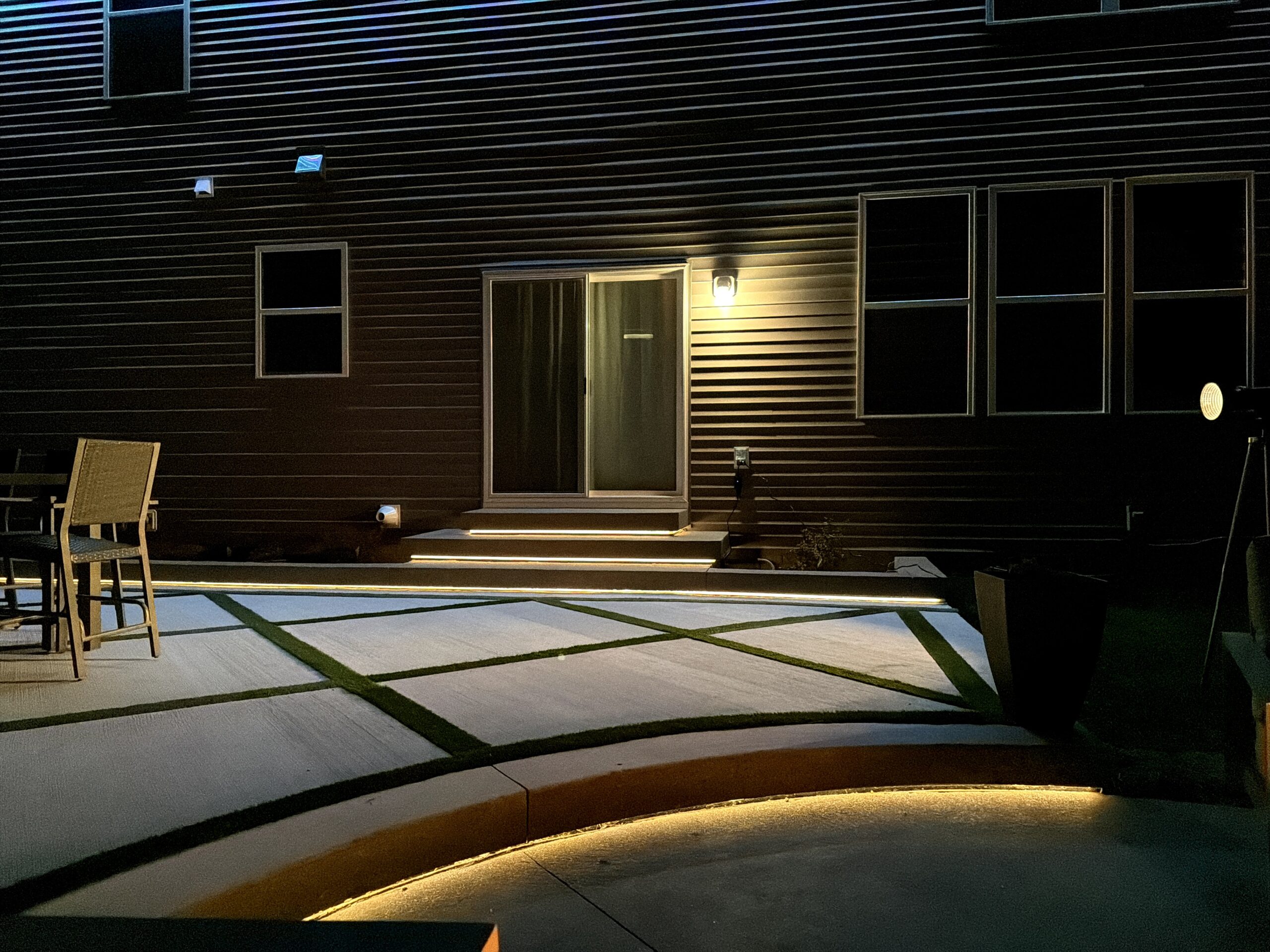 Night view of a modern house exterior with dark siding, lit doorway and a row of windows along the wall; patio seating visible on the left and illuminated stepping stones leading to the door.