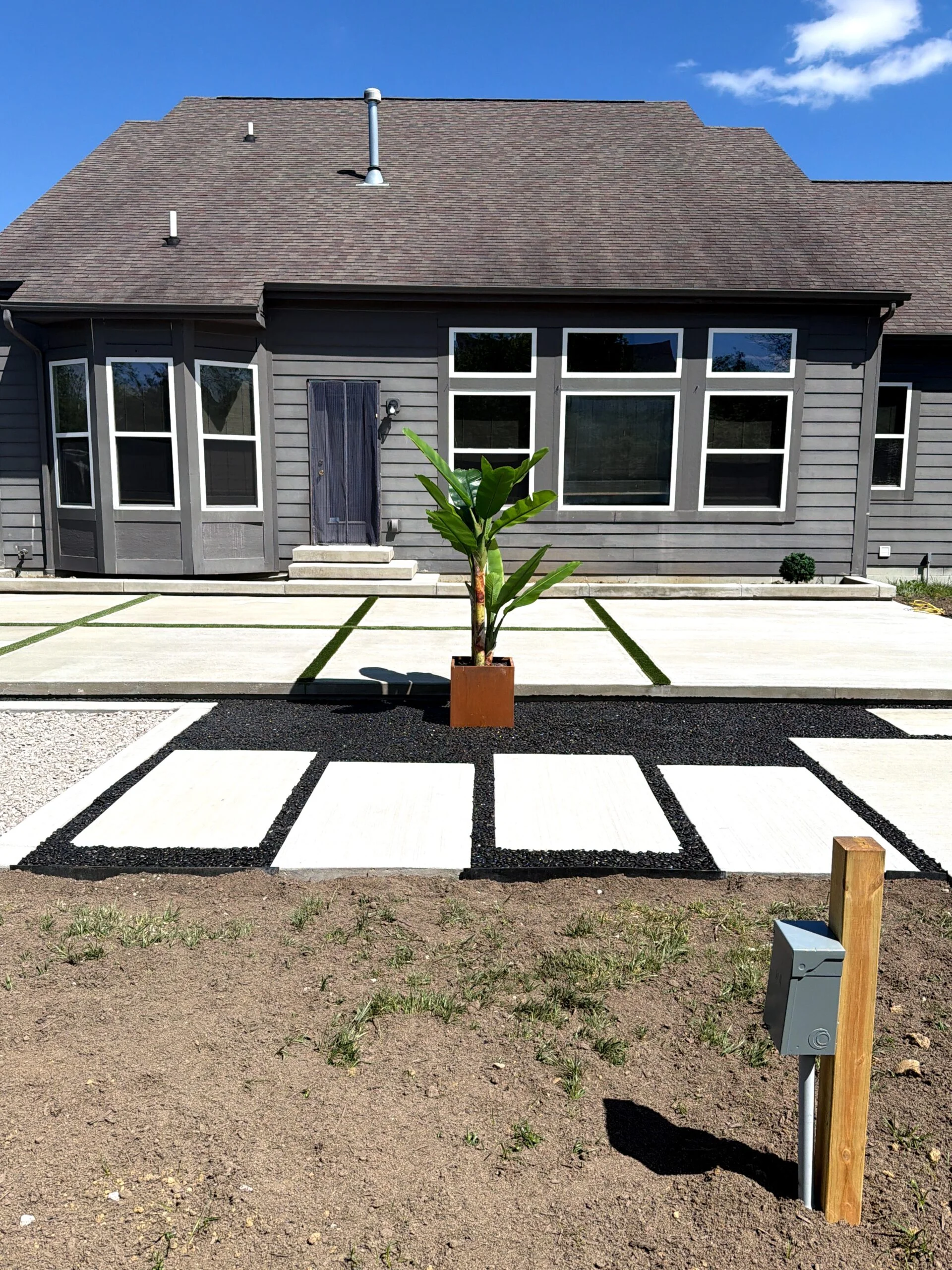 Front of a gray modern house with large white-trimmed windows; a potted banana plant sits on a geometric concrete-and-gravel driveway in front of the home.