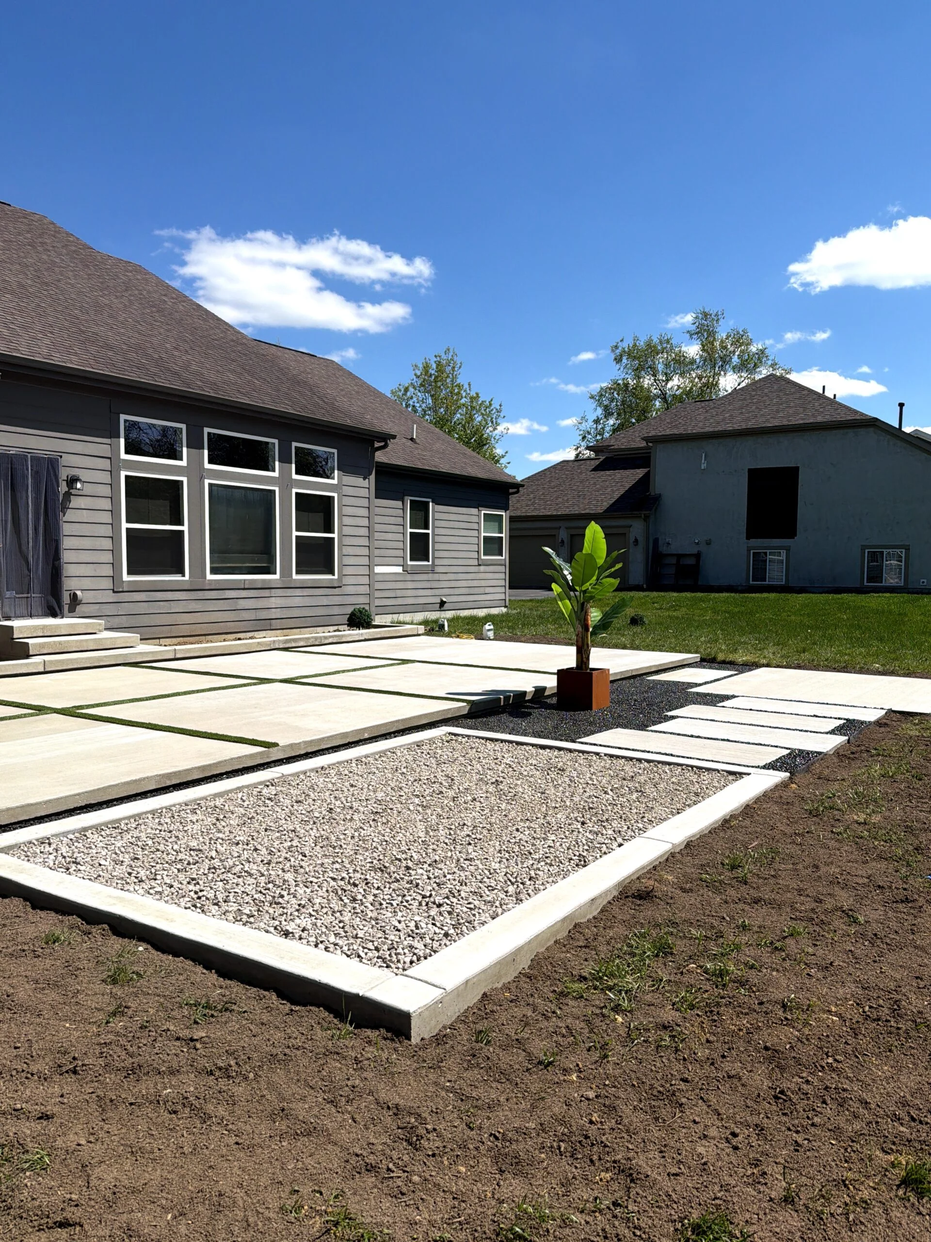 Modern gray house with large white-trimmed windows, concrete stepping stones, and a potted banana plant in the yard under a blue sky.