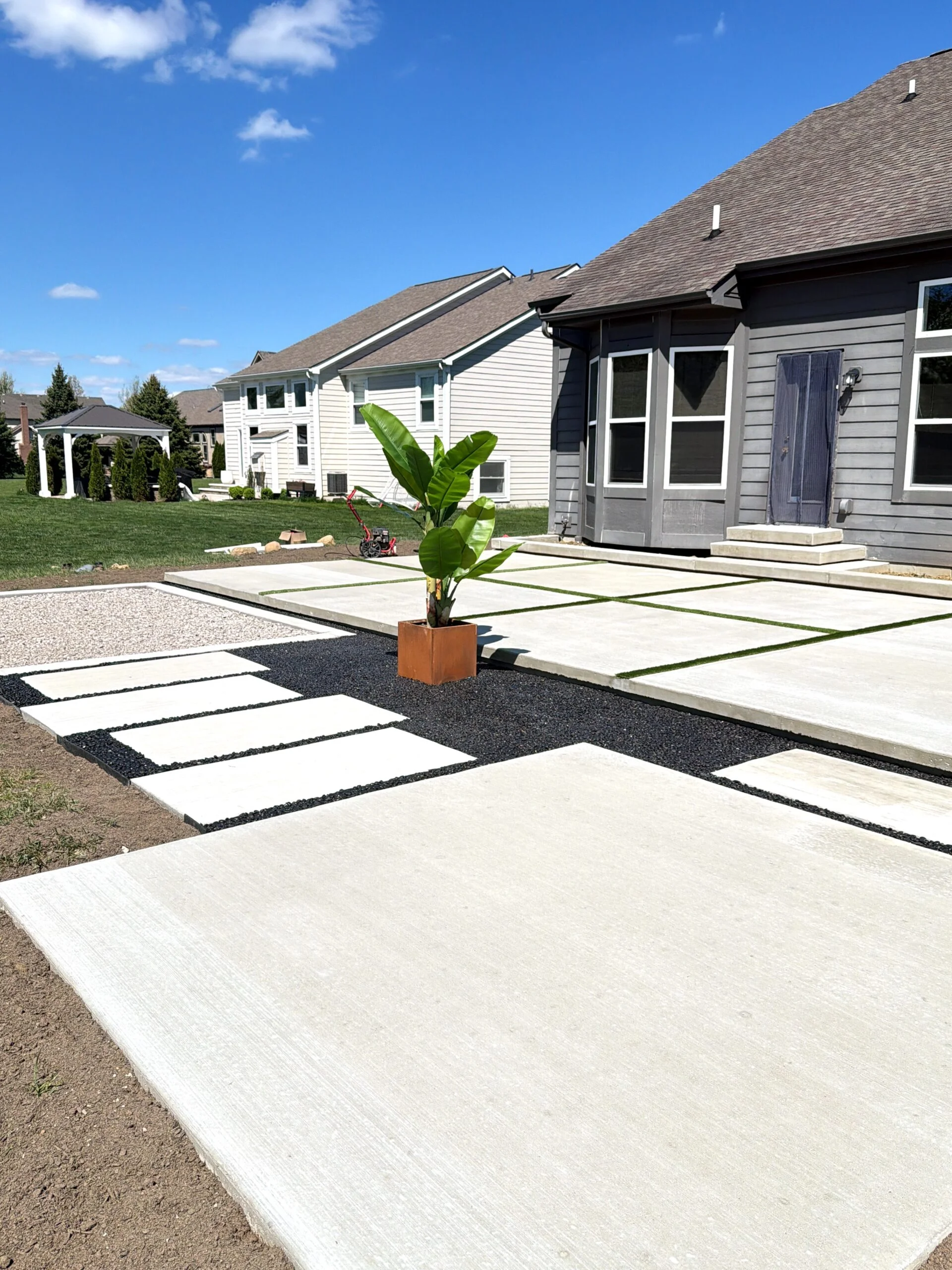 Suburban front yards with modern houses and a geometric concrete stepping-stone walkway leading to the gray house; a potted banana plant sits in the foreground under a blue sky.