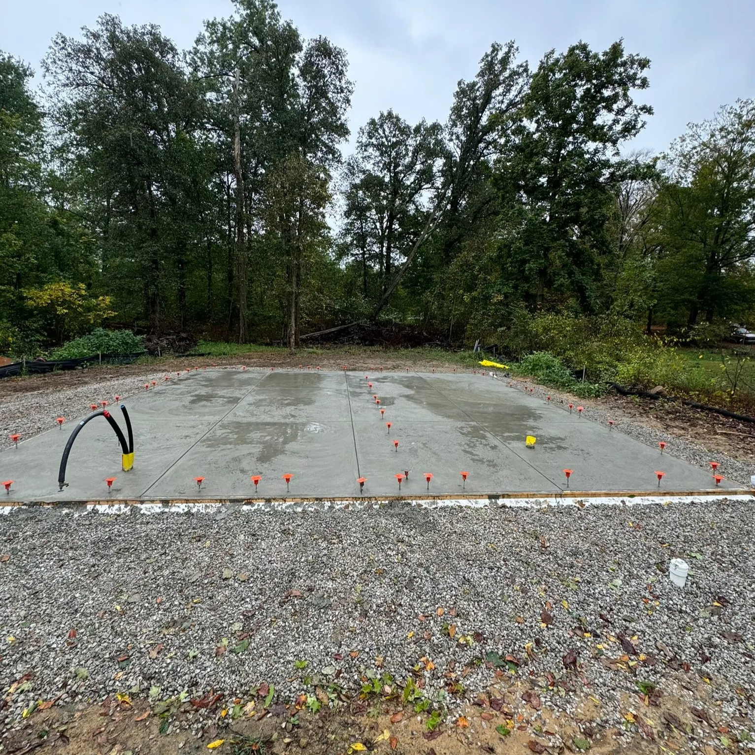 Fresh concrete slab foundation on a gravel lot with orange edge markers and a hose nearby, trees in the background.