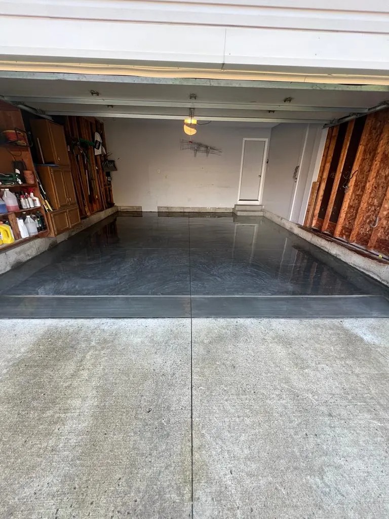 Polished epoxy-finished garage floor with a central seam, shelves on the left, and a back wall with a door in a residential garage.