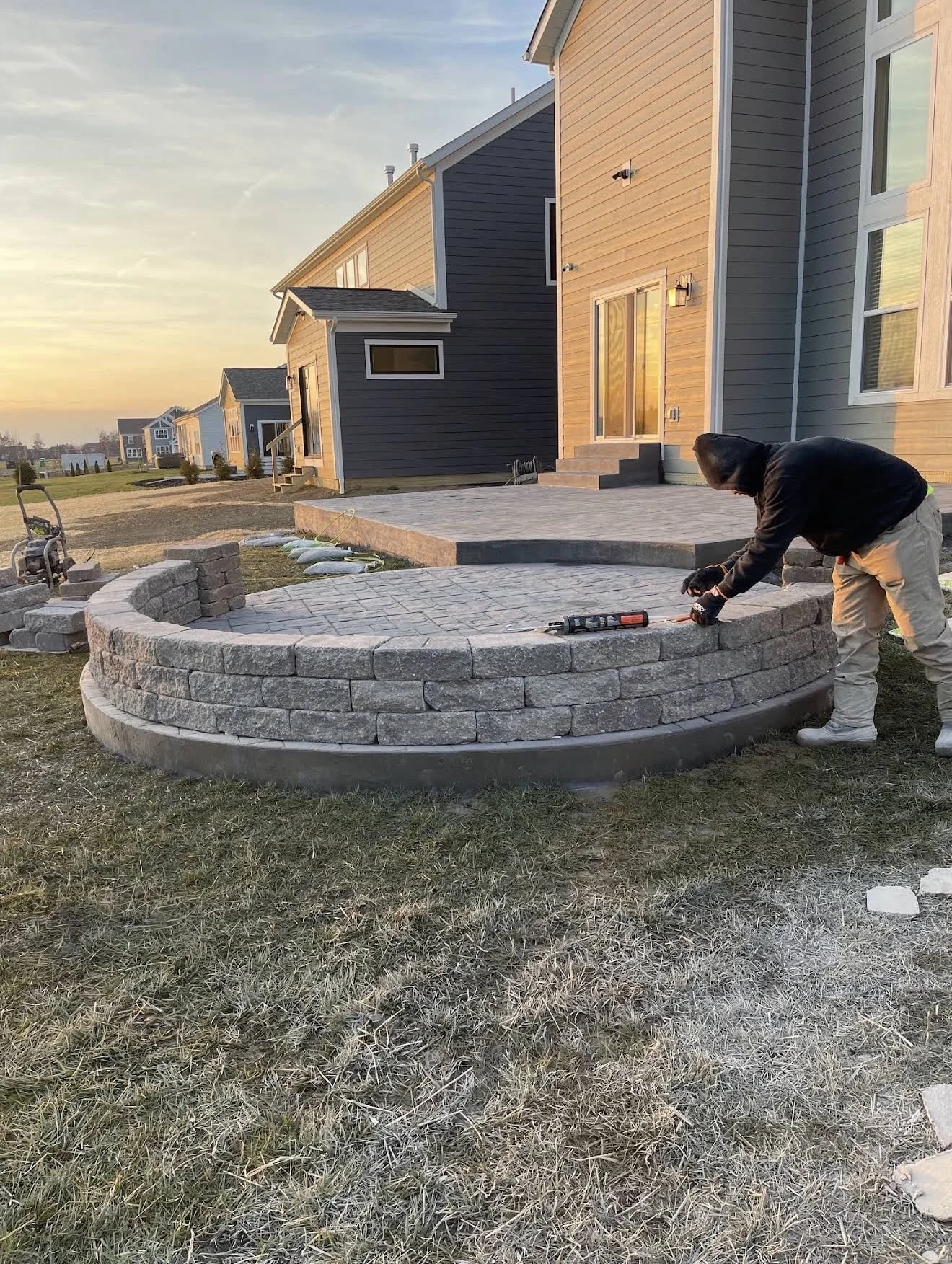 Worker lays curved stacked-stone edging around a paved patio in front of a modern blue-gray house at sunset/late afternoon light.